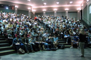 blonde professor stands on the floor of a lecture hall and gives a lecture to over 200 students piloting